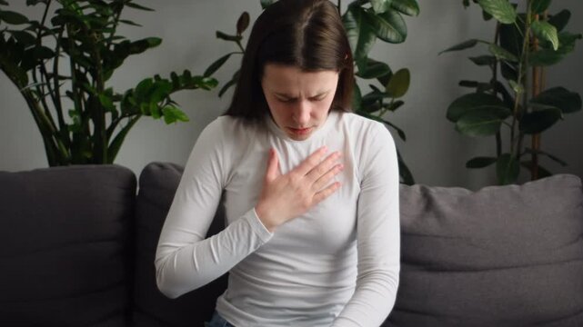 Unhealthy young caucasian woman having severe chest pain while sitting alone on sofa. Sad worried female suffers from heartache at home, infarction or heart disease concept