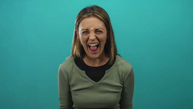 Young blonde woman shouting with mouth open and tongue out in turquoise studio; anger release catharsis.