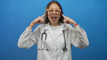 Naklejka premium Woman doctor in white coat with stethoscope presses fingers to ears and shouts in blue studio; stress fatigue.