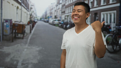 Fototapeta premium Hispanic young man pointing thumb over shoulder on street while smiling; casual confidence happiness.