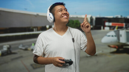 Young hispanic man with headset holds game controller and points finger upward in airport terminal;...