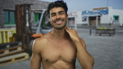 Shirtless man with muscular chest smiles and scratches head next to a forklift in front of a...