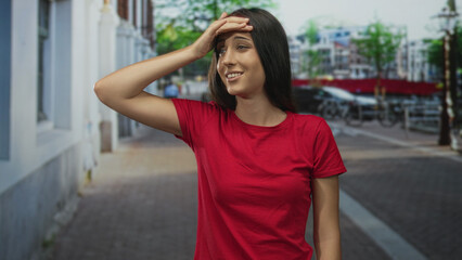 Fototapeta premium Young hispanic woman in red shirt with hand on forehead on a city street, palm to brow gesture; embarrassment.