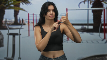 Young hispanic woman makes phone gesture with hand to ear in outdoor gymnasium with fitness...