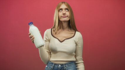 Redhead woman holds a white milk bottle in one hand, puckering lips and rolling eyes while wearing a cream sweater and jeans in a studio red backdrop; skepticism taste testing.