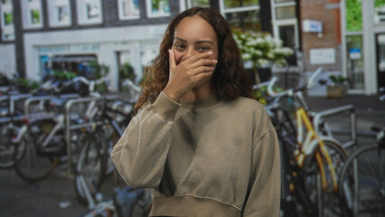Obraz premium Woman covering mouth with hand on a busy amsterdam street lined with bicycles and a storefront, wearing beige sweatshirt; amusement.