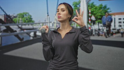 Obraz premium Woman holding dumbbell at shoulder, hand gripping weight, face visible on street; strength confidence focus.