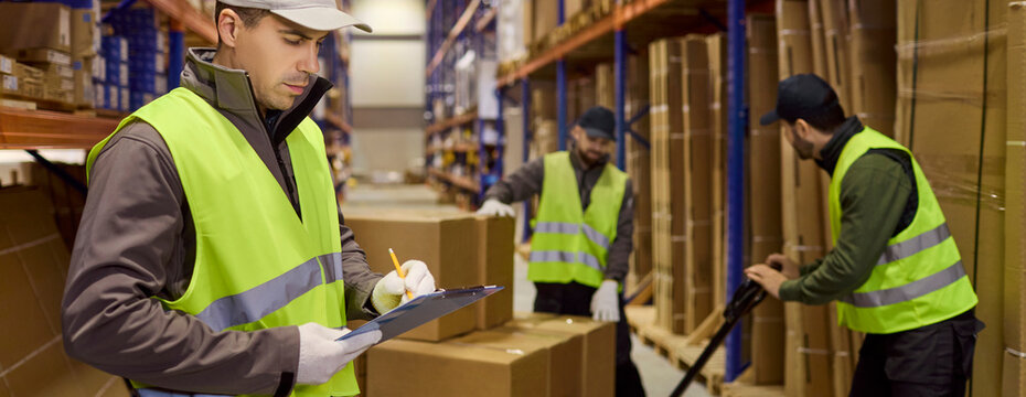 Warehouse worker checks inventory on clipboard. In a busy warehouse, a loader moves boxes on a pallet while staff track shipping orders. Concept shows efficient logistics tracking process.