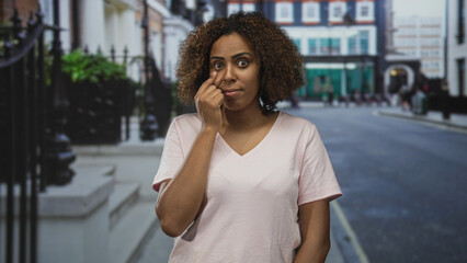 Fototapeta premium Woman in pale pink shirt points finger to lower eyelid in a city street outside shops; health concern.