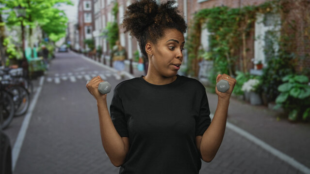 Woman holds dumbbells and lifts light weights on a city street with a wry smirk and small ring visible; playful determination.