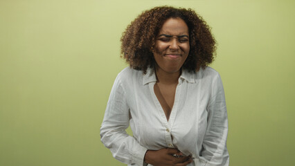Obraz premium Young african american woman with curly hair grimacing and holding her stomach while wearing a white shirt in a pale green studio; nausea.