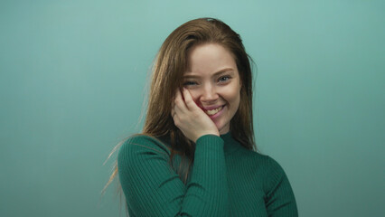 Fototapeta premium Woman wearing green sweater placing hand on cheek and smiling with closed eyes in teal studio setting; amusement.
