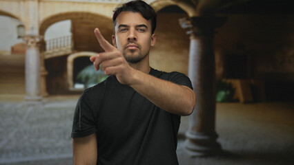 Young man in black shirt stands on old town street pointing forward with confident expression under outdoor arches. © Krakenimages.com