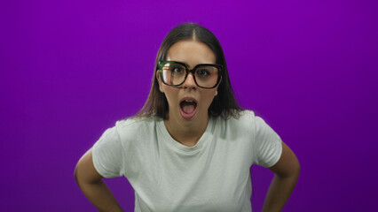 Young brunette woman wearing oversized glasses with mouth open gasping and hands on hips in studio with purple backdrop; surprise curiosity.