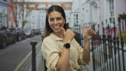 Woman, young brunette, thumbs pointing to self showing smartwatch on wrist on a city street lined with pastel townhouses and parked cars; confidence celebration.