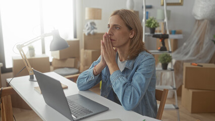 Obraz premium Woman at desk with laptop, hands pressed to mouth in building amid moving boxes, ladder and drill, looking pensive and concentrated; moving anxiety reflection.