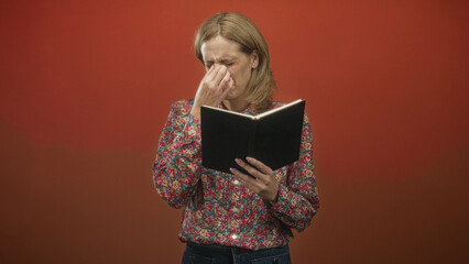 Woman in floral blouse with blonde hair reads a black notebook while pinches her nose, frowns and squints in studio; disgust concentration.