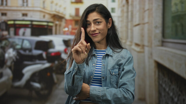 Woman points index finger up on street with denim jacket and striped top, arm folded across chest; wry skepticism.