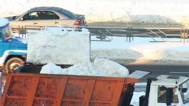 Orange front loader bucket full of fresh white snow unloading into large rusty orange dump truck bed street maintenance during cold winter day city road cleanup