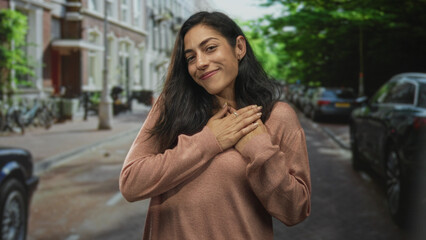 Fototapeta premium Young hispanic woman clasping hands to chest on a residential street with parked cars and bicycles; gratitude warmth.