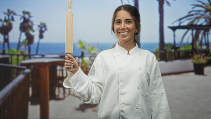 Woman chef holds rolling pin upright on a building terrace overlooking the ocean, smiling directly at camera while wearing a white chef coat  confidence skill. © Krakenimages.com