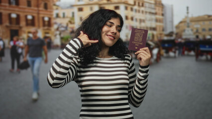 Fototapeta premium Woman holding passport and making call gesture in street near historic building, smiling and posing with striped top; travel pride.