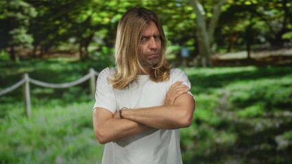Obraz premium Blond man with long hair crosses bare arms in forest wearing white shirt with furrowed brow; skepticism doubt.