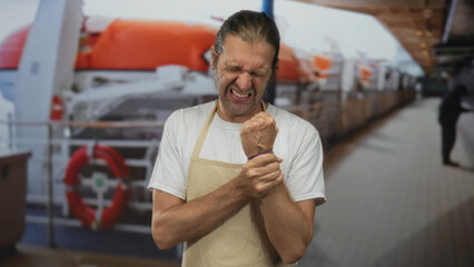 Fototapeta premium Blond man with long hair in apron clutches his right wrist and grimaces inside building; discomfort.