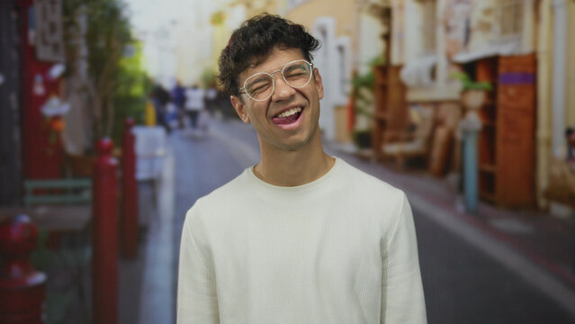 Man wearing glasses with tongue out winking in colorful urban street scene with blurred pedestrians; playful mood.