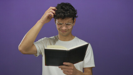 Hispanic man wearing glasses scratching head while reading book in purple studio; confusion...