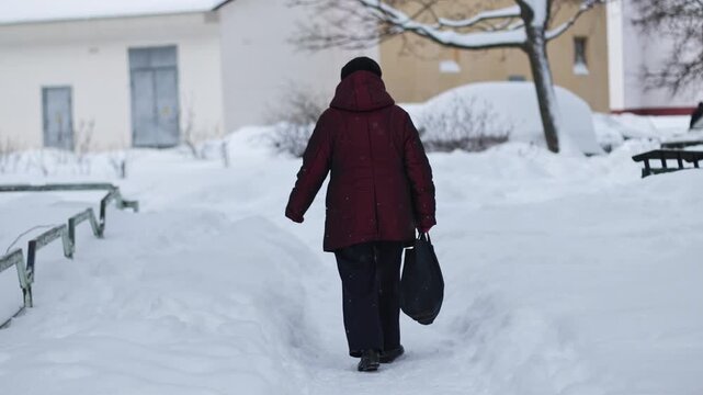 Rear view of older adult woman walking on path covered with fresh white snow. Woman wears dark red winter coat and dark pants, carries black bag. Buildings and tree present. Slow motion x4