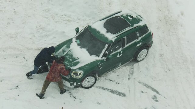 Overhead perspective of two individuals manually pushing green sport utility vehicle stranded in heavy snow. Vehicle wheels spinning, people struggle in severe winter weather.