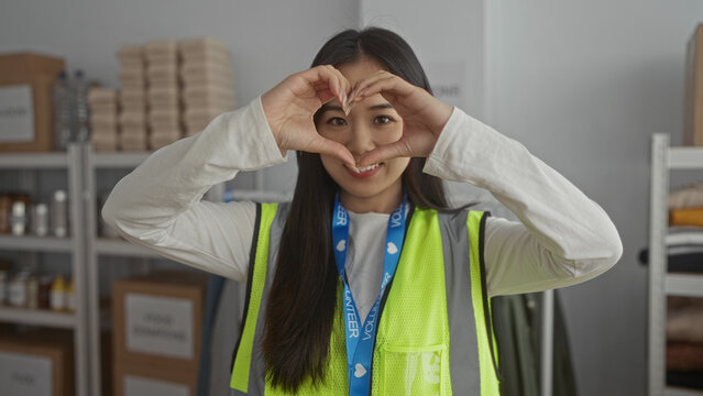 Young woman in a reflective vest makes a heart gesture at a volunteer center, surrounded by charity supplies, emphasizing community and kindness.