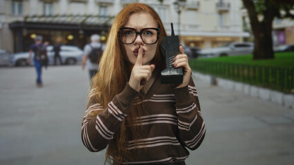 Fototapeta premium Redhead woman holding a brick phone, finger to lips for silence on a street with storefronts and parked cars; secrecy caution.