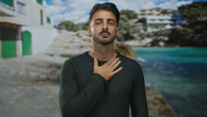 Fototapeta premium Man with short beard and bare ear studs places hand on chest in front of a coastal building with green shutters; sincerity gratitude.