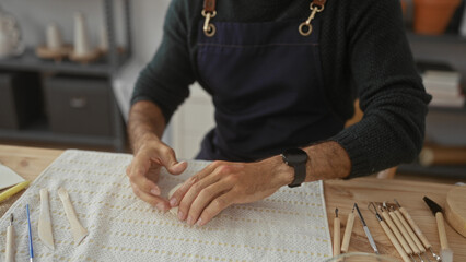 Man kneads clay with bare hands over a wooden worktable surrounded by pottery tools in studio; creativity.