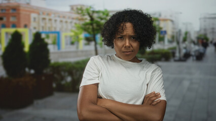 Obraz premium Woman wearing white t shirt with arms crossed on city sidewalk near brick building and planters; doubt.