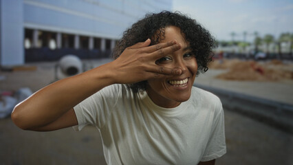 Fototapeta premium Woman wearing white shirt peeks through fingers with smiling eyes at building under construction in sunshine; joy.