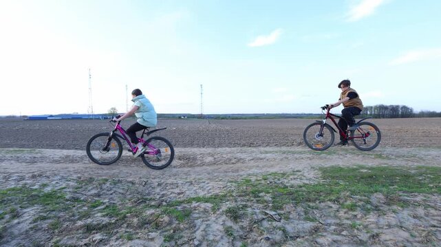 Two teens ride bikes side by side along a rural dirt track, plowed fields stretch wide, sky is clear, and motion conveys joyful, carefree youth in open countryside.