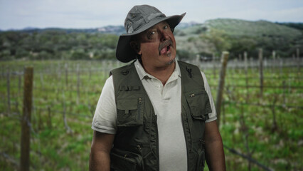 Fototapeta premium Man fisherman tilts head and sticks tongue out wearing wide brim hat and fishing vest in a vineyard, hands relaxed; playful curiosity.