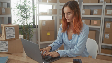 Redhead woman holding smartphone and typing on laptop in building, surrounded by shipping boxes and...