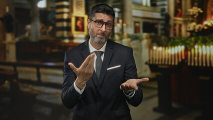 Fototapeta premium Man in dark suit and tie gestures with palms up and hand to chest inside ornate church building with candles and pews; pleading faith.