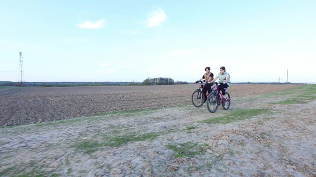 Two teens ride bikes side by side along a rural dirt track, plowed fields stretch wide, sky is clear, and motion conveys joyful, carefree youth in open countryside.