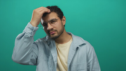 Fototapeta premium Young hispanic man wearing glasses in casual shirt touches head with hand in studio on green backdrop; confusion self reflection.