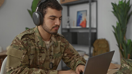 Fototapeta premium Man in camouflage uniform wearing headphones and dog tags typing on laptop in living room; focused discipline.