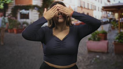 Fototapeta premium Woman plus size shielding eyes with hand on cobblestone street, smiling with nose ring and exposed midriff, casual daytime scene; carefree joy.
