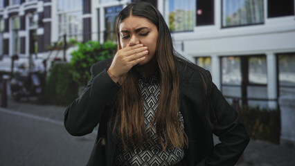 Fototapeta premium Woman holding hand to forehead on street in front of building windows, eyes closed and grimacing; stress fatigue.
