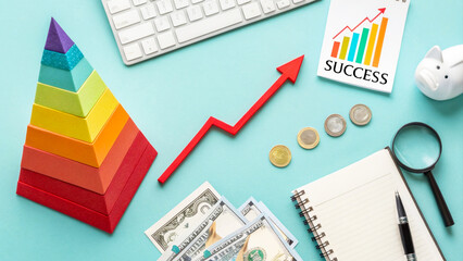 Obraz premium Flat lay of financial planning items: colorful pyramid, red upward arrow, keyboard, money, coins, notebook, magnifier and success chart on blue background.