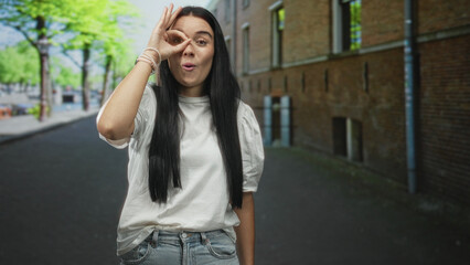 Fototapeta premium Young hispanic woman standing on a city street by a brick building, making ok sign with hand over eye while smiling in a white t shirt; playful curiosity.