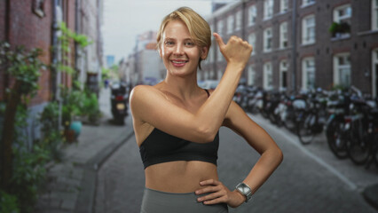 Fototapeta premium Woman holding fist to left shoulder with other hand on hip, showing midriff and wearing smartwatch while smiling in street lined with bicycles and brick buildings; confidence vitality wellness.